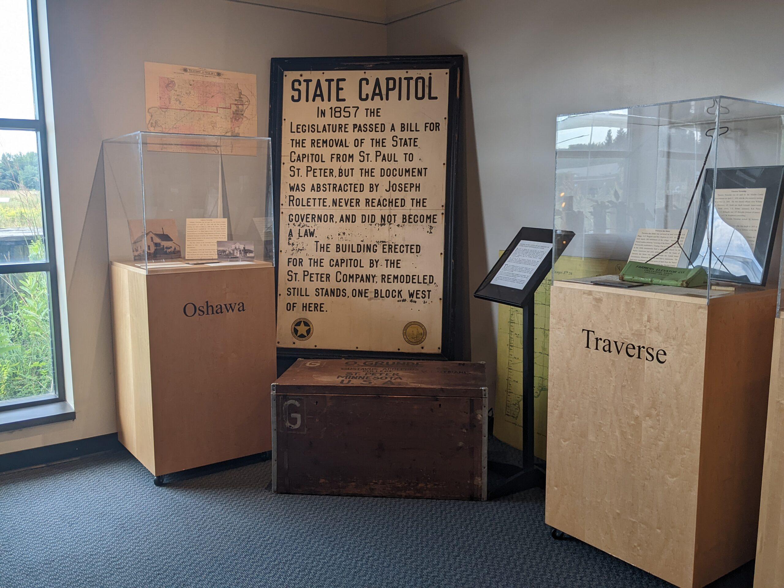 Historical exhibit on the 1857 State Capitol removal, featuring informational displays and artifacts, including old documents under glass cases, in a museum-like setting with natural lighting.