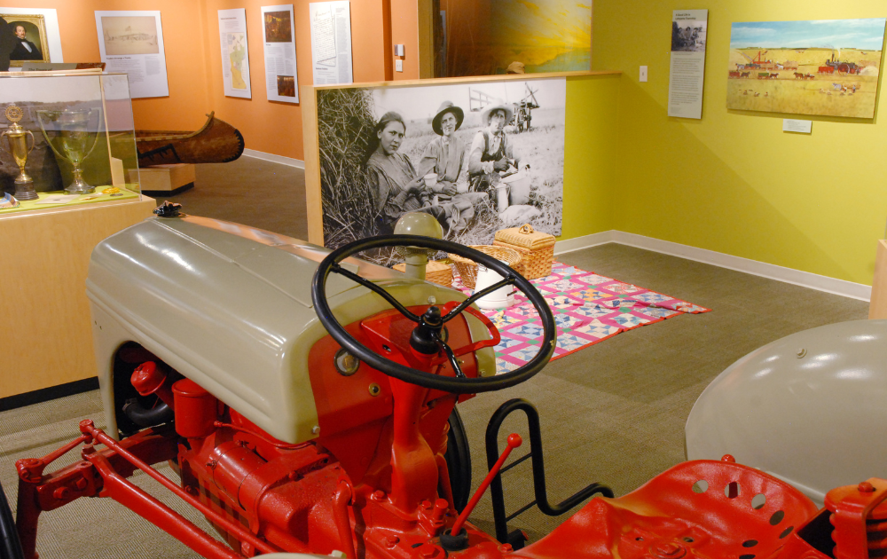 Vintage red tractor on display at a historical museum exhibition with large black-and-white and colorful paintings reflecting agricultural history on the walls.