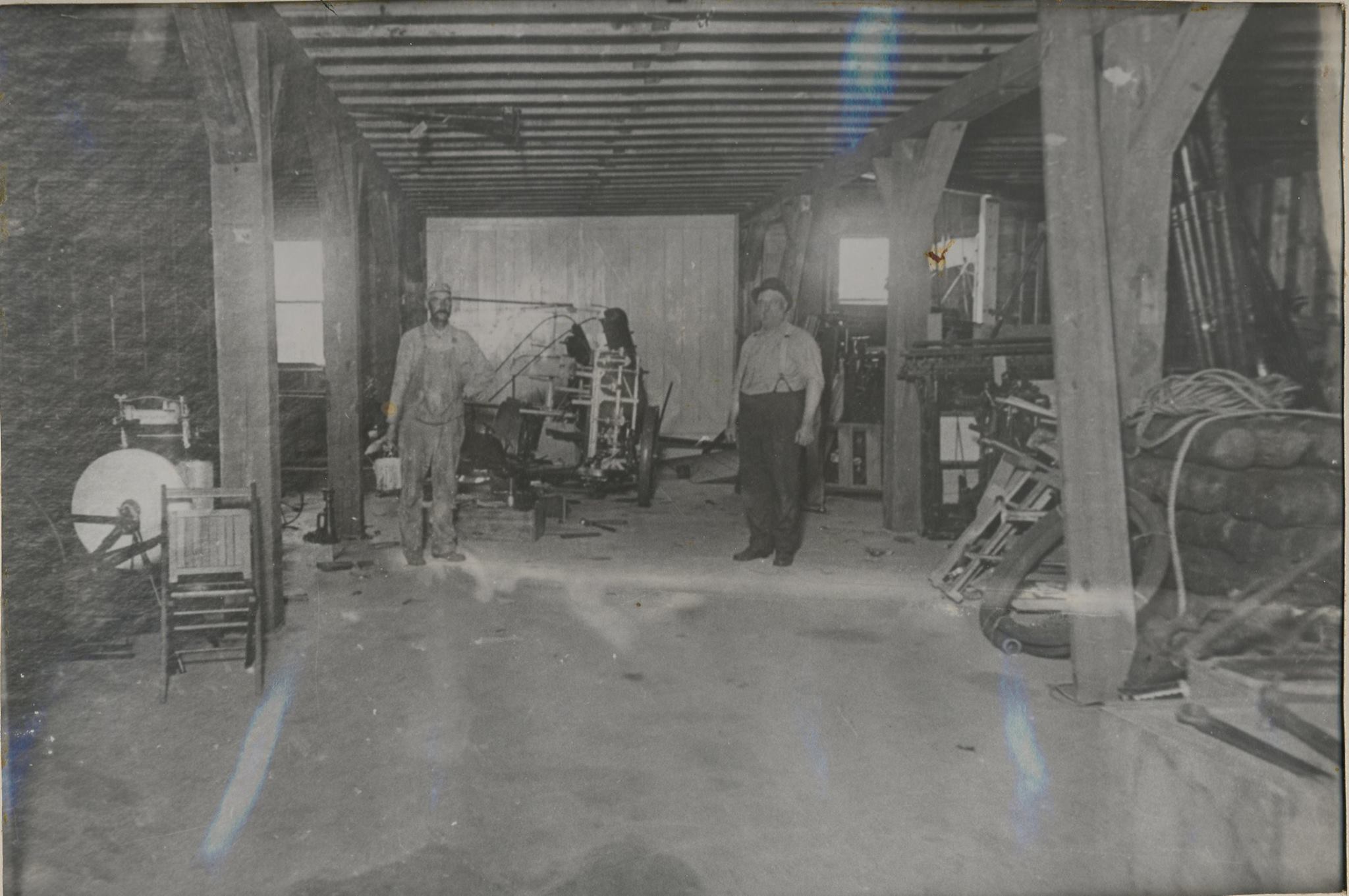 Two men stand in a large, cluttered workshop with wooden beams running across the ceiling. The workshop contains various tools and equipment, including a large machine in the middle and stacks of materials on the right. The men wear work clothes and hats.