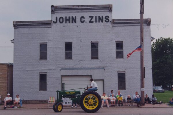 A person drives a vintage green tractor in front of a large, gray building labeled "JOHN C. ZINS." An American flag hangs on a pole to the right. A group of people, including children and adults, are sitting on folding chairs along the sidewalk, watching the scene.