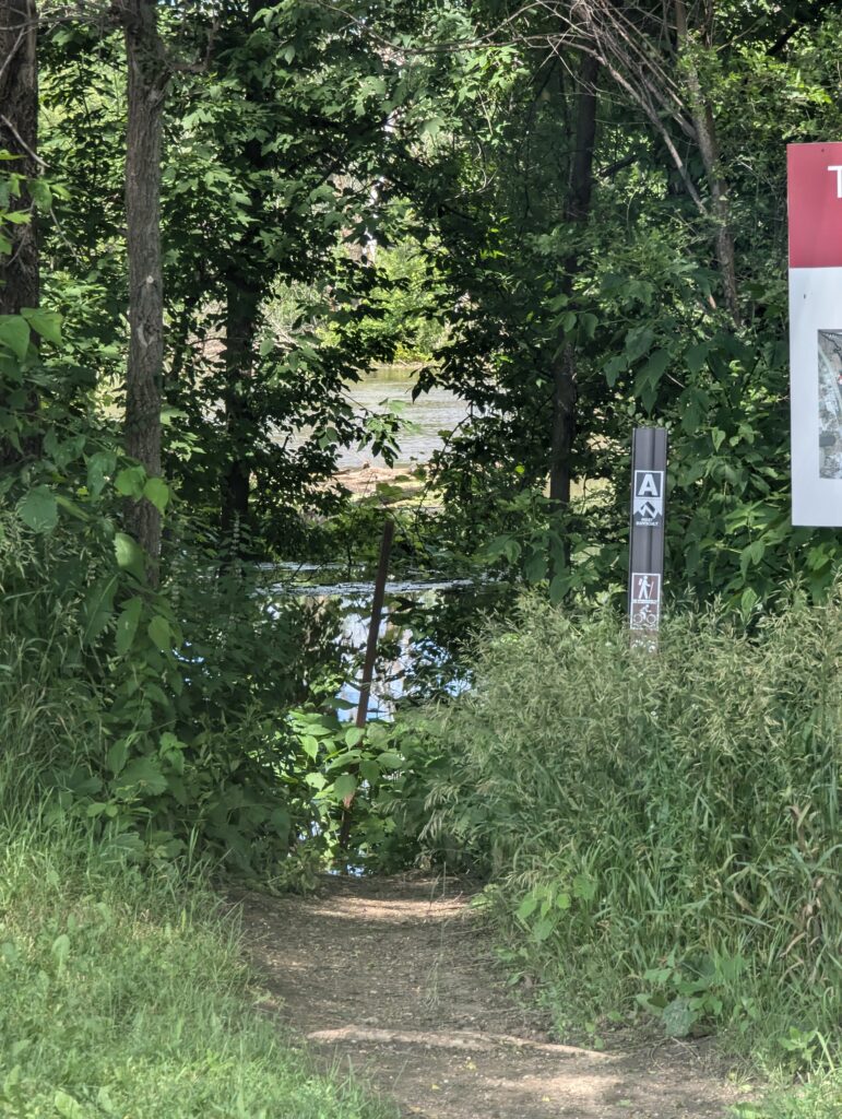 A narrow dirt path leads through dense greenery toward a body of water partially visible in the background. Trees and bushes surround the pathway. A post with a sign and a red board with a partial map are on the right side of the path.