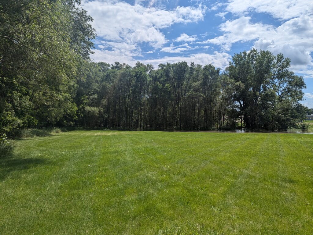 A lush, green field with trimmed grass extends towards a cluster of tall, leafy trees under a partly cloudy blue sky. The sun shines through, casting light and shadows on the peaceful, open landscape.