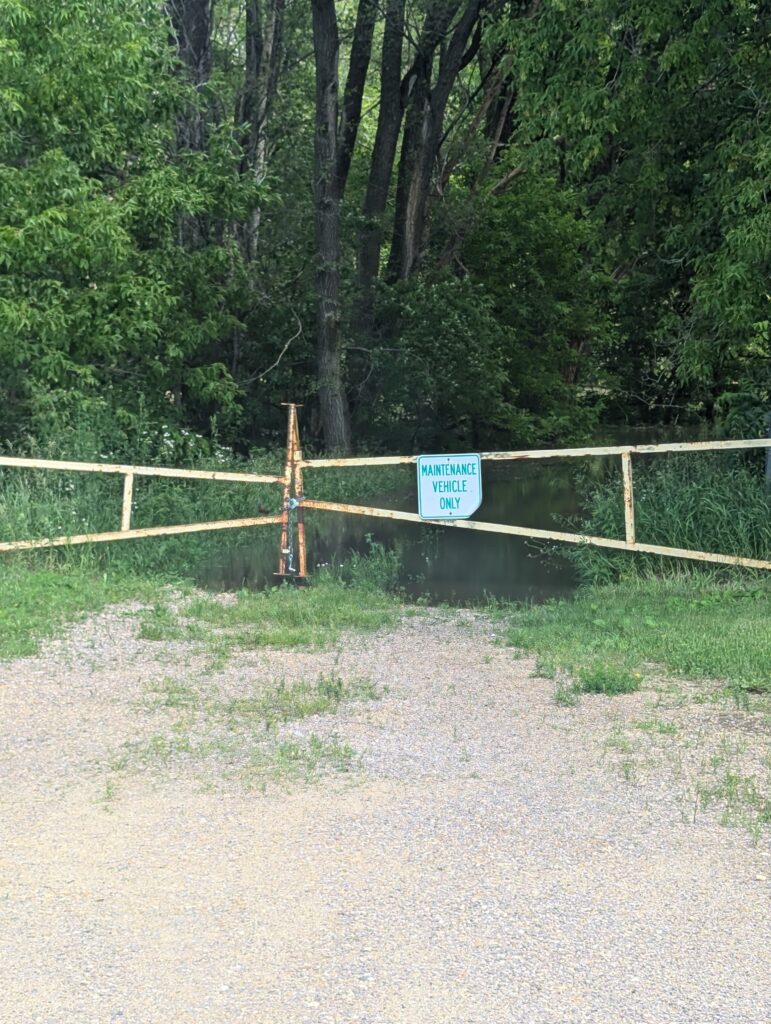 A yellow metal gate blocks a gravel path leading into a forested area with dense greenery. The gate has a sign that reads "Maintenance Vehicles Only." Grass surrounds the gravel path, and tall trees are visible in the background.