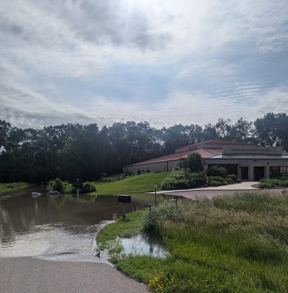 A low building labeled "Forest Arts Education Center" stands under a partly cloudy sky. A flooded area covers parts of the driveway and adjacent grassy area. In the background, dense trees surround the center, casting shadows on the scene.