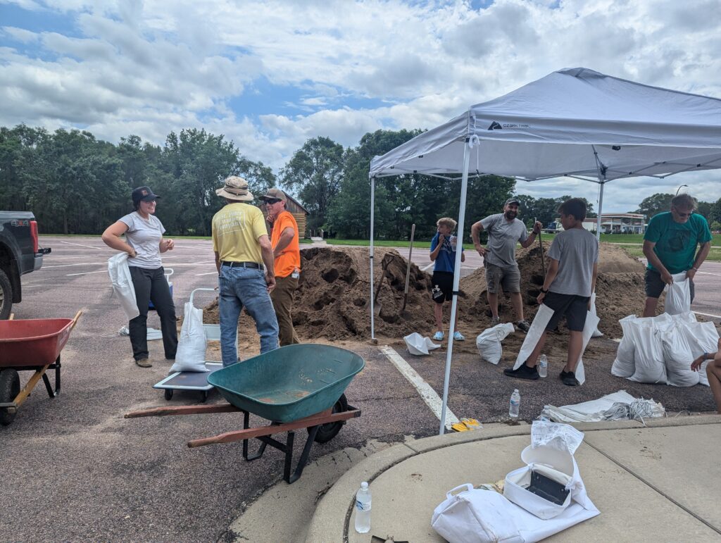 A group of people stands around a large mound of dirt, filling sandbags under a white canopy tent in a parking lot. Wheelbarrows, shovels, and sandbags are scattered around. The sky is partly cloudy, and trees line the background.