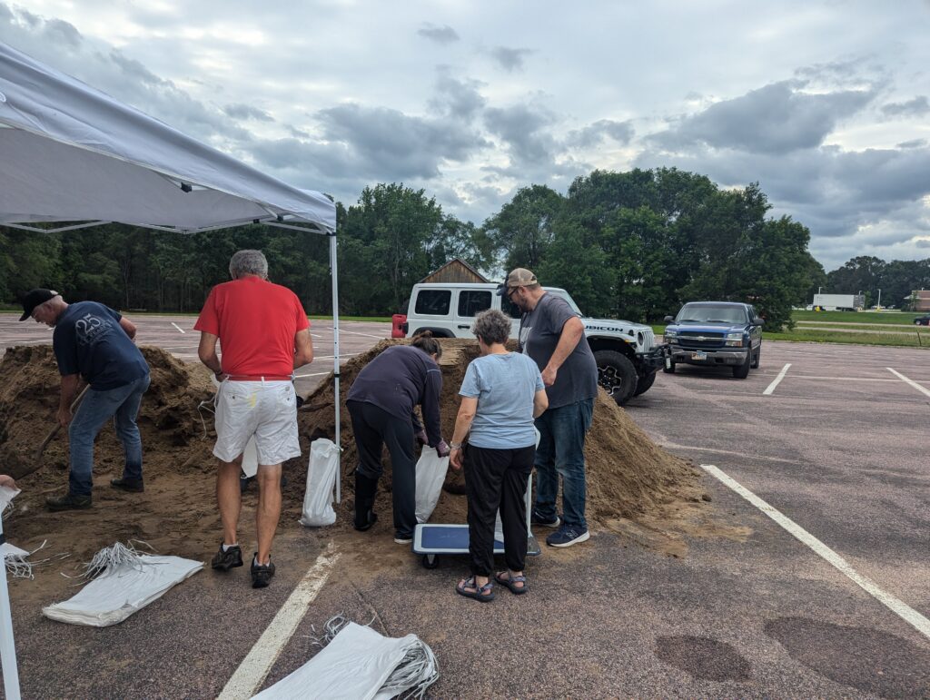 People are filling sandbags from a pile of sand in a parking lot. A white vehicle and a blue truck are parked nearby. The sky is overcast, and there are trees in the background. Several filled sandbags and empty sacks are on the ground.