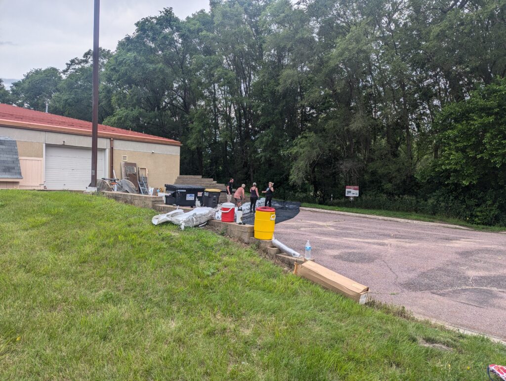 People are gathered near various construction materials and equipment on a driveway beside a building with a garage door. Trees are in the background. The scene appears to involve some form of outdoor work or construction.