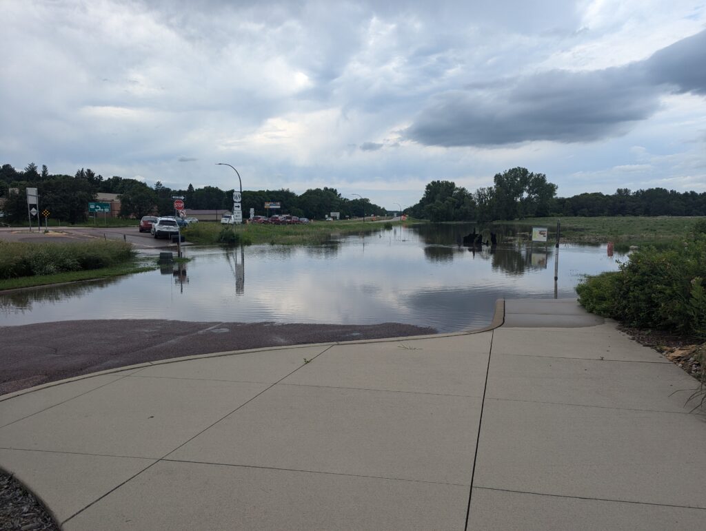 A flooded roadway, with water covering the pavement and extending onto the sidewalk. Traffic signs are partially submerged, and cars are parked on the dry part of the road in the distance. Trees and greenery surround the area under a cloudy sky.