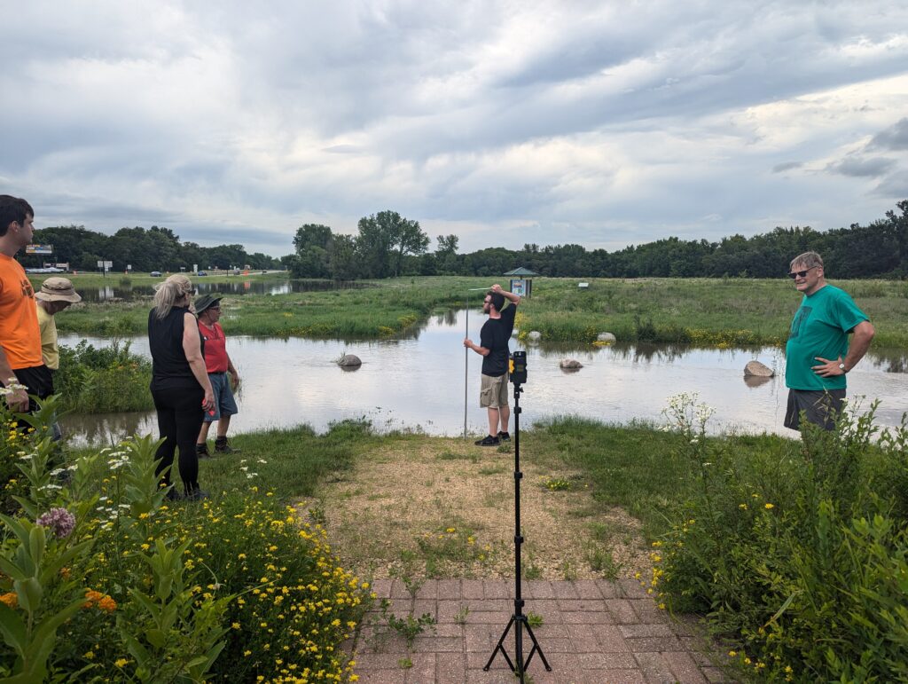 Five people stand near a body of water surrounded by greenery under a cloudy sky. One person holds a measuring tool, while another stands by a tripod-mounted equipment. The setting appears to be a park or nature reserve with flowers in the foreground.