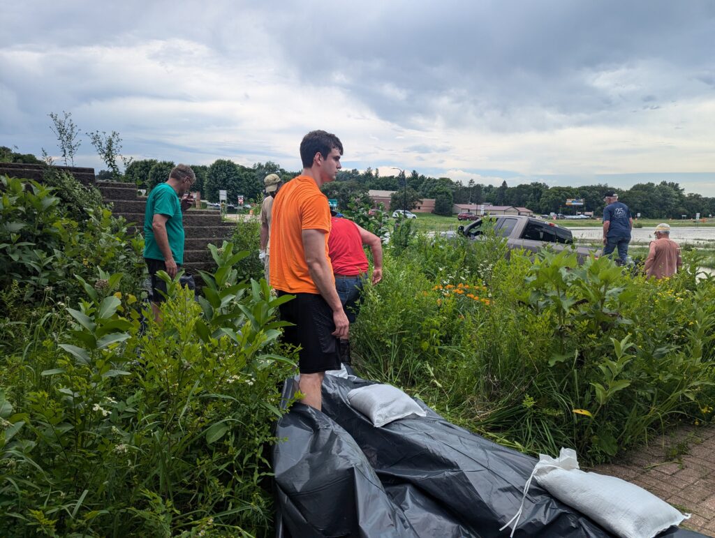 A group of people stands amidst overgrown vegetation near a partially submerged vehicle. One person in an orange shirt is in the foreground, while others work together in the background under a cloudy sky. Sandbags are visible on the ground.