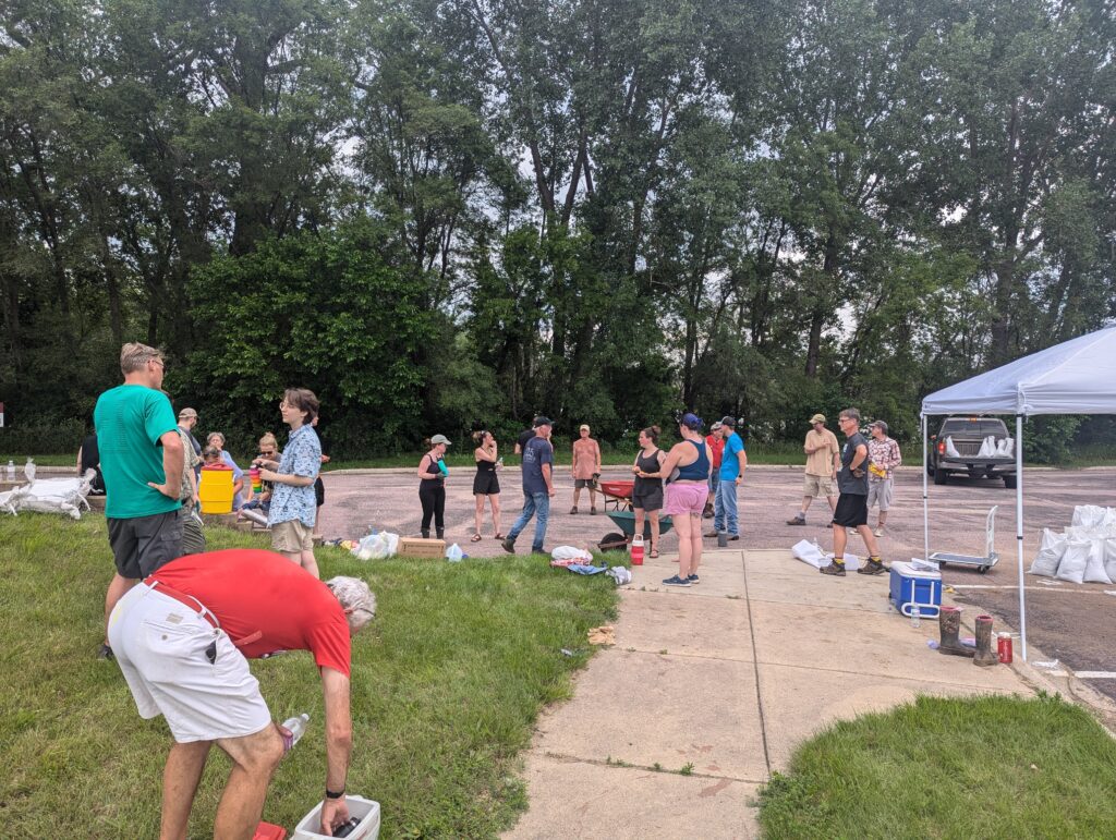 A group of people gathers outdoors near a wooded area on a concrete path and grassy field. Some are bending over plastic bins, while others stand and converse. A canopy tent is set up, and bags are scattered around, suggesting an organized outdoor event.