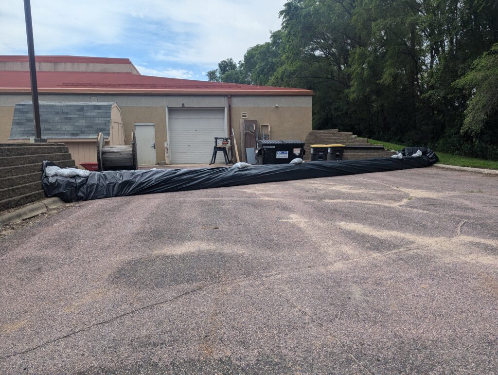 A parking lot with a long black tarp covering an object stretched across it. Behind the tarp, there is a small storage shed, a beige building with a large garage door, and a few dumpsters. The background has trees and a partly cloudy sky.