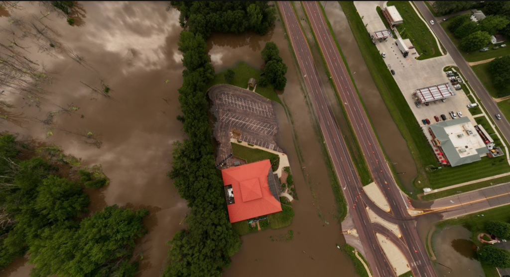 Aerial view of severe flooding with water submerging a large area, including a building with a red roof, adjacent parking lot, and nearby roads. Trees surround the flooded area, which is next to a highway. Buildings and a parking area are visible on the right.