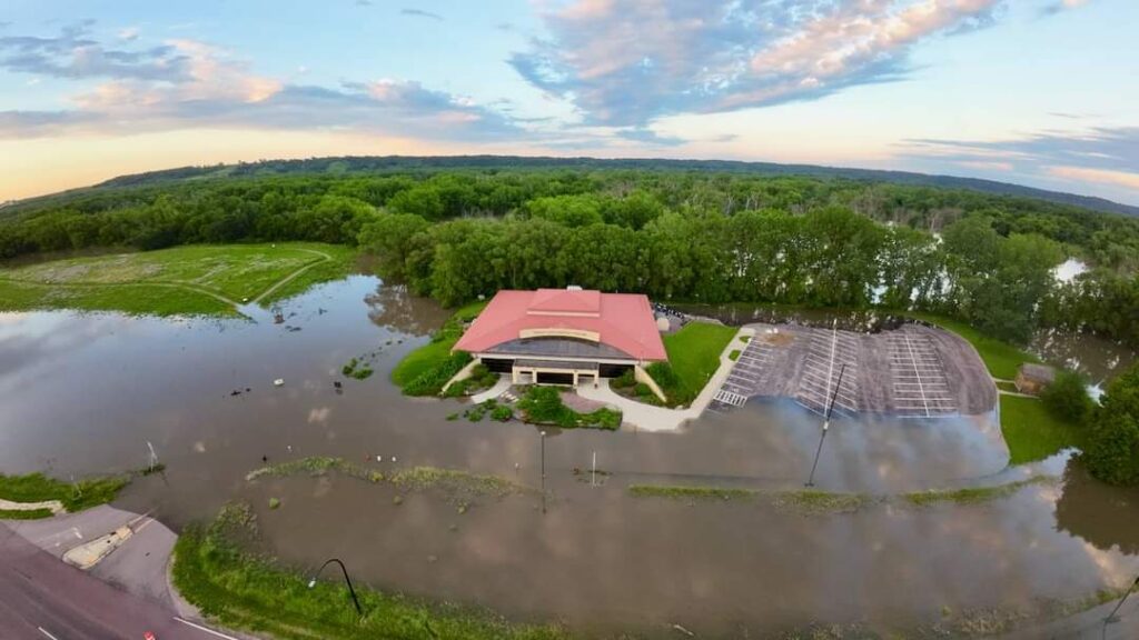 Aerial view of a building with an orange roof surrounded by extensive flooding. The adjacent parking lot and surrounding greenery are partially submerged in water. Trees and partially flooded fields can be seen in the background under a partly cloudy sky.