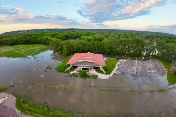 Aerial view of a building with an orange roof surrounded by extensive flooding. The adjacent parking lot and surrounding greenery are partially submerged in water. Trees and partially flooded fields can be seen in the background under a partly cloudy sky.