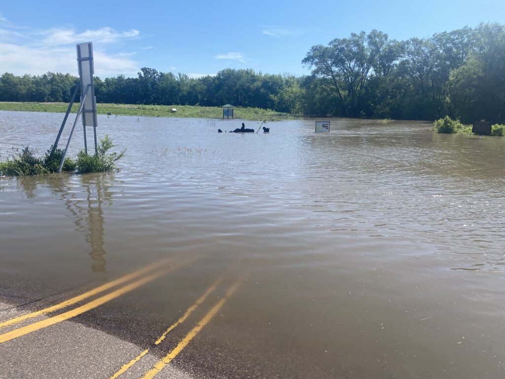 A flooded roadway is submerged under murky water, with road signs barely visible above the surface. Trees and greenery surround the area, reflecting on the water. The sky is clear with scattered clouds, hinting at a recent storm or heavy rainfall.