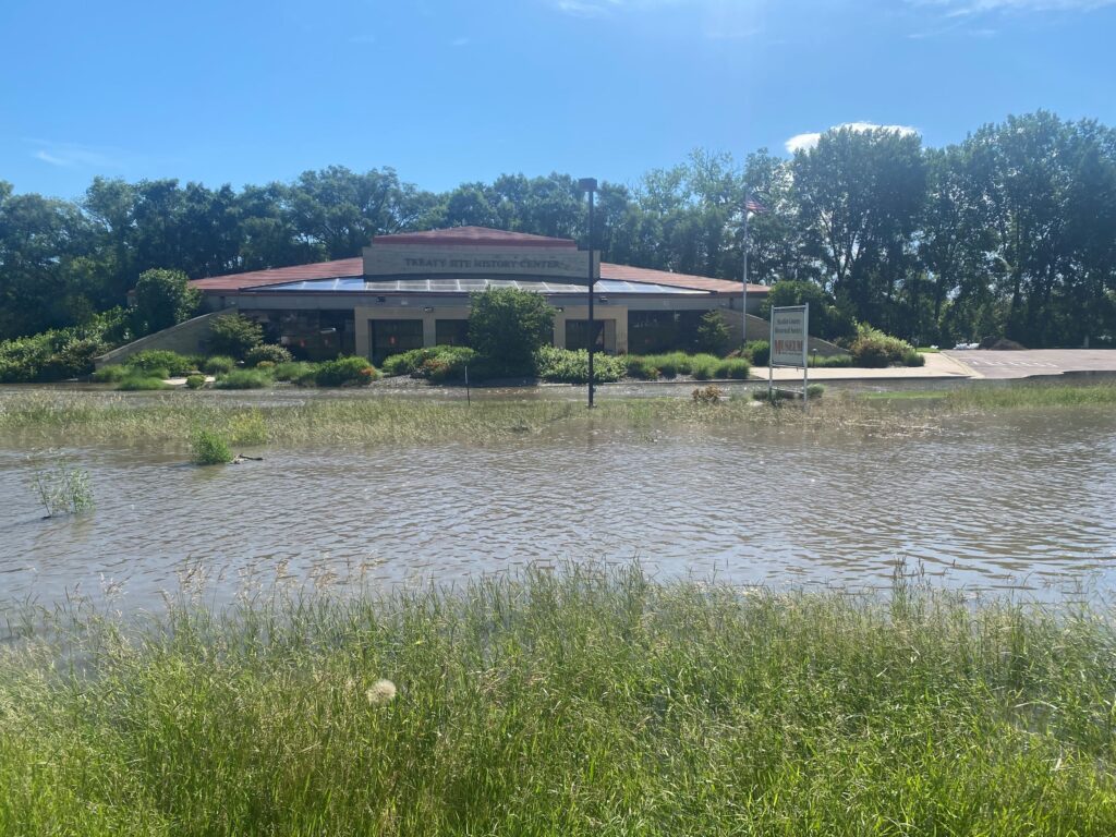 A building with a red roof identified as "Tri-County Restaurant & Catering" is surrounded by floodwater. The foreground shows waterlogged grass and a partially submerged parking lot. Trees and a clear blue sky are visible in the background.