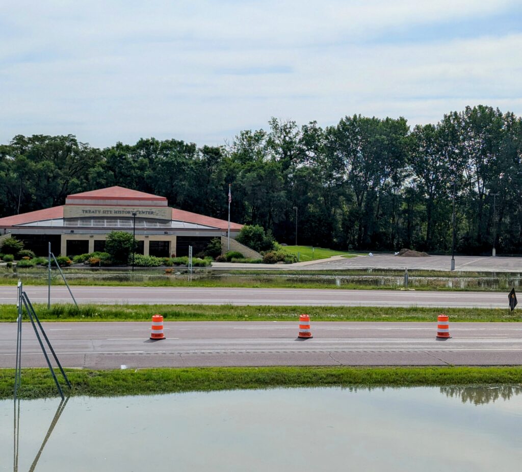 The image shows the Illinois State Police District 6 Headquarters building in the background, with trees behind it. In front, there are multiple traffic cones along a road, and a flooded area is visible in the foreground on a cloudy day.