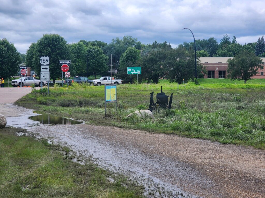 A rural roadside with a stop sign at an intersection. There are direction signs indicating routes 169 and nearby towns. A muddy path and a small map display are in the foreground. Trees and a building are visible in the background under a cloudy sky.