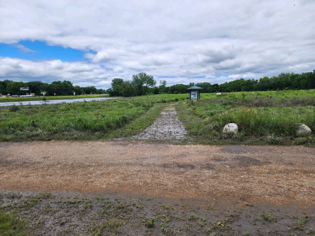 A dirt and gravel path extends through a green, grassy field with scattered trees in the distance under a partly cloudy sky. The path intersects a small gravel road, leading towards a blue information sign posted on the right.