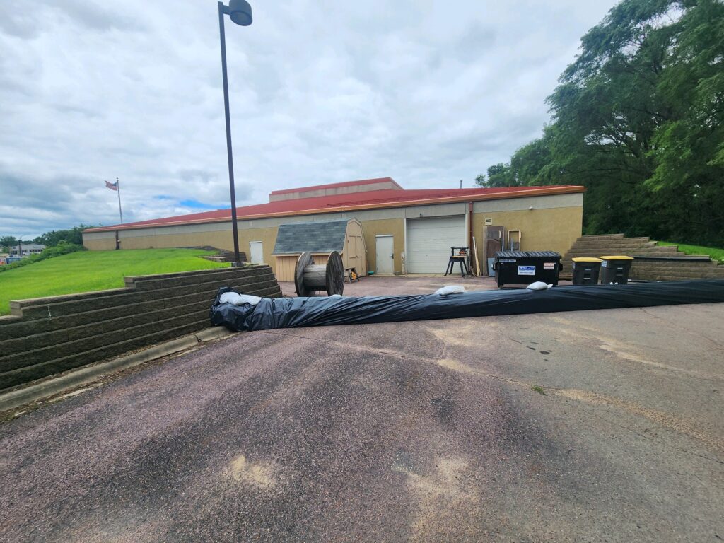 A large plastic sheet covers and extends from a cylindrical object towards the corner of a tan warehouse, blocking access to a garage door. The area is surrounded by concrete and grassy slopes with trees in the background and a cloudy sky above. .