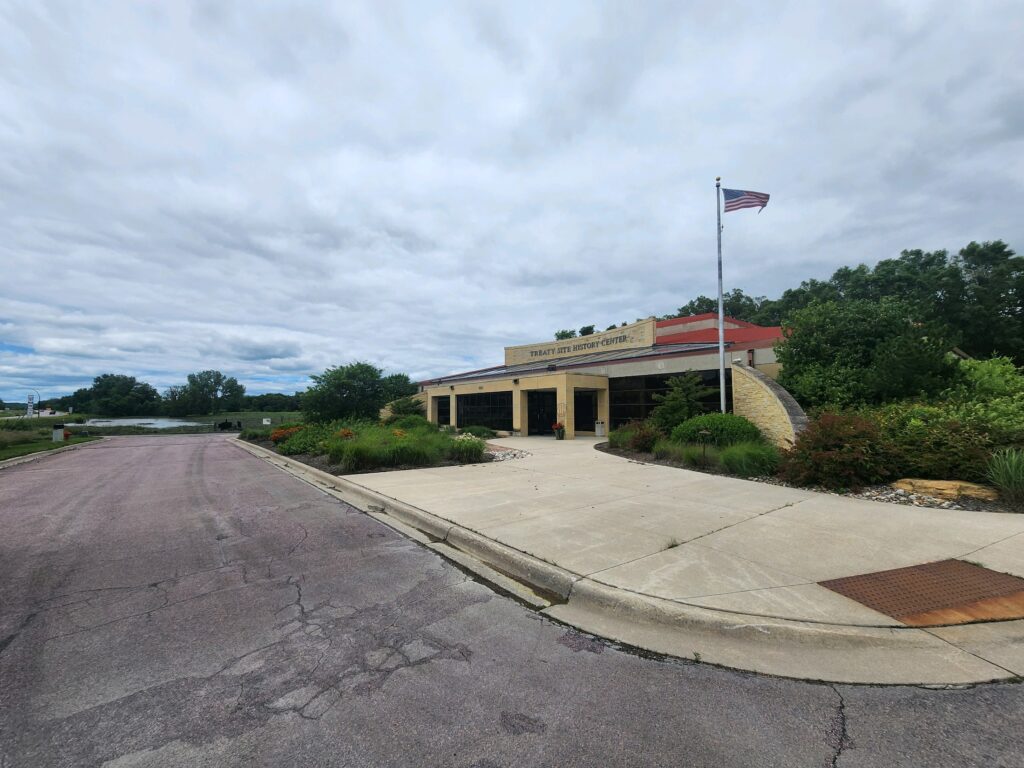 A building with a sign that reads "Tracy Area High School" is situated near a road with an American flag flying on a pole outside. The structure is surrounded by greenery and bushes, with a mostly cloudy sky overhead.