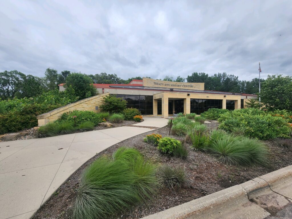 Front view of the Treaty Site History Center, surrounded by green shrubbery and plants, with a paved walkway leading to the entrance. The building has a red and beige facade with large windows. The sky is cloudy and overcast.