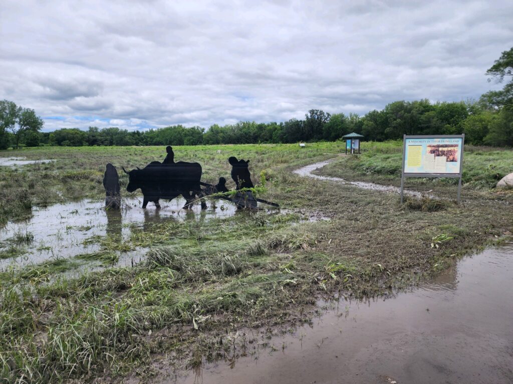 A flooded outdoor pathway with standing water, a cut-out silhouette installation of farm animals and people, informational signs, and green trees in the background. The sky is overcast with gray clouds.