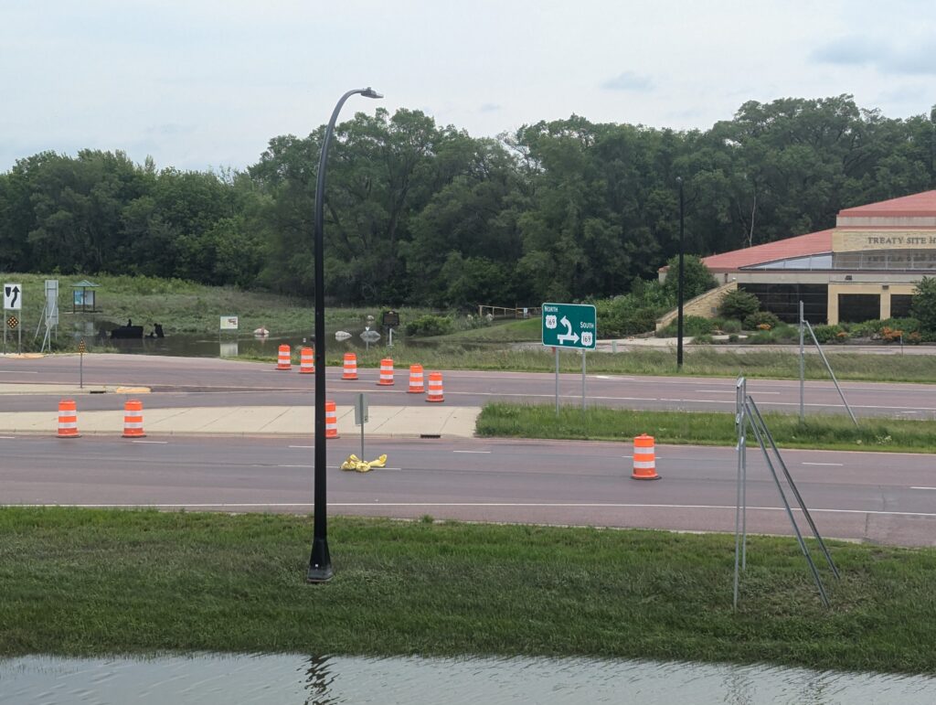 A road closed with orange barrels lined up and a 'Keep Right' sign indicating a detour. A street light is viewable on the left, and a large green sign is on the right. There is a small grassy area and some buildings in the background.