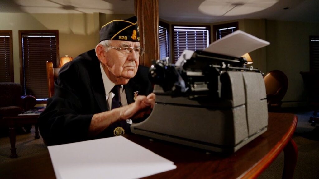 An elderly man in a military uniform and cap sits at a typewriter on a wooden table, surrounded by papers. The room has several windows with partially closed blinds and warm lighting from lamps.