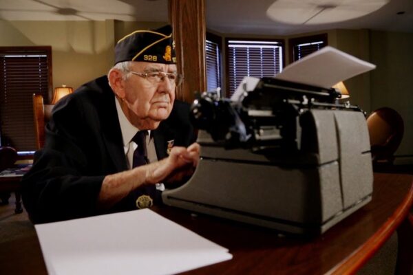 An elderly man in a military uniform and cap sits at a typewriter on a wooden table, surrounded by papers. The room has several windows with partially closed blinds and warm lighting from lamps.